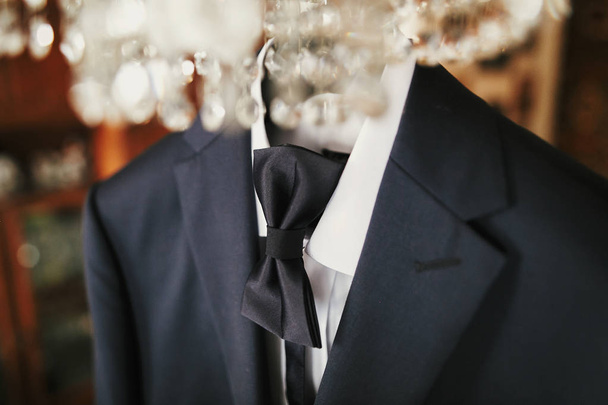 stylish black suit with white shirt and bow tie on hanger on luxury chandelier. morning preparations before wedding day. groom outfit - Photo, Image