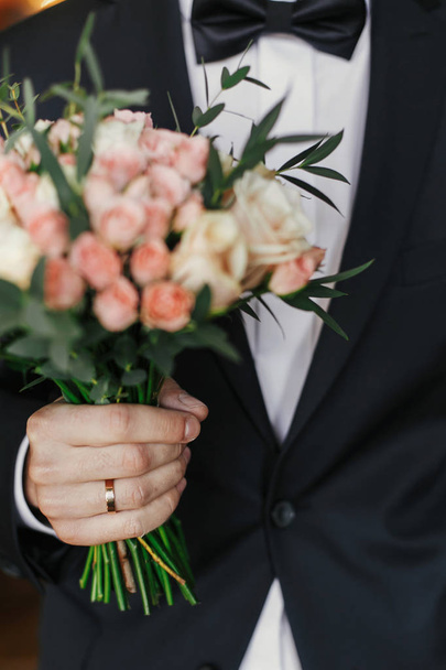 Жених держит стильный букет с розами и эвкалиптом. утренняя подготовка перед свадьбой. Жених ждет свою невесту
 - Фото, изображение