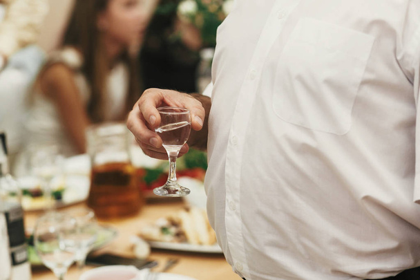 Mann hält Glas Wodka und stößt auf Hochzeitsempfang an. Menschen jubeln und stoßen bei Festessen im Restaurant mit Getränken an - Foto, Bild