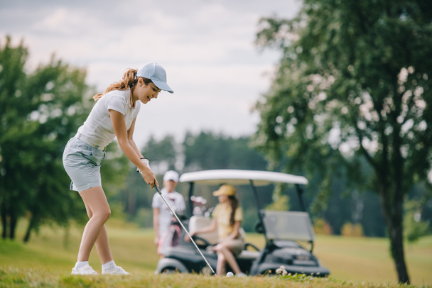 foco seletivo da mulher com clube de golfe jogando golfe e amigos descansando no carrinho de golfe no gramado verde
 - Foto, Imagem