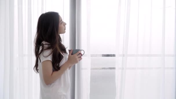 junge asiatische Frau genießt ihren Kaffee oder Tee am nassen Fenster in ihrem Schlafzimmer zu Hause. schöne Teenager weiblich trinken Getränke und entspannen Sie sich an regnerischen Tagen. - Filmmaterial, Video