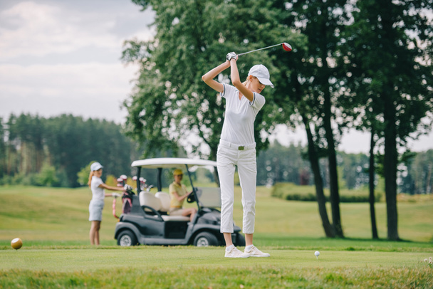 Selektiver Fokus der Frau mit Golfschläger beim Golfspielen und Freundinnen beim Ausruhen am Golfcart auf grünem Rasen - Foto, Bild