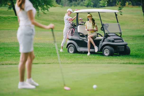 selectieve aandacht van vrouw met golfclub en vrienden rustend op de golfkar op groen gazon - Foto, afbeelding