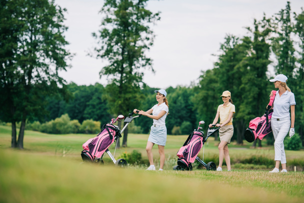 smiling women in caps with golf equipment walking on golf course - Valokuva, kuva
