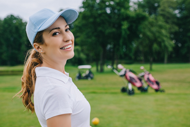 vista lateral da mulher sorridente no pólo e boné olhando para a câmera no campo de golfe
 - Foto, Imagem