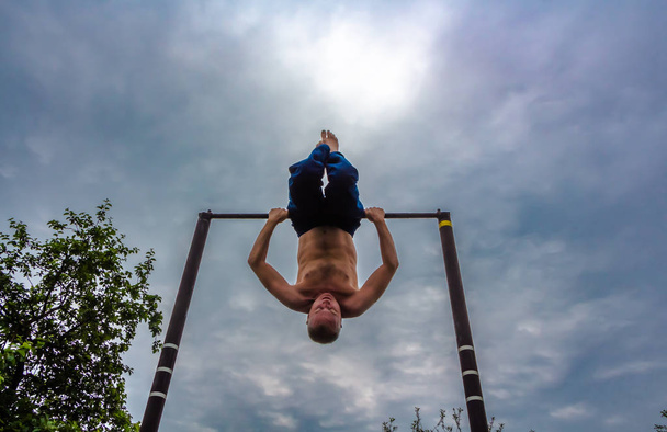 Muž s holé torzo v modrých kalhotách vzhůru nohama dělá jeho ranní cvičení na vodorovný pruh - Fotografie, Obrázek
