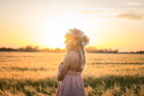 beautiful blonde lady in head flower wreath during sunrise in summer wheat fields - Fotó, kép