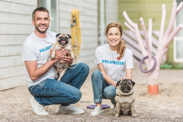 two volunteers of animals shelter squatting with pug dogs and looking at camera - Photo, Image