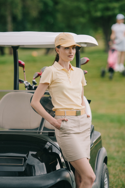 foco seletivo de jogador de golfe feminino em boné com as mãos nos bolsos no carrinho de golfe no gramado verde
 - Foto, Imagem