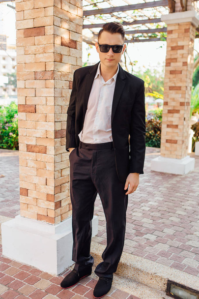 Young stylish handsome man posing in modern city park, wearing suit and sunglasses. - Zdjęcie, obraz