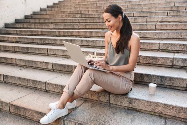 Photo of caucasian beautiful woman 20s wearing casual summer outfit sitting on street stairs with takeaway coffee and typing on silver laptop - Фото, зображення