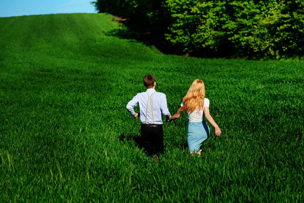 Young happy lovers running on meadow with green grass and blue sky, From the back. - Zdjęcie, obraz
