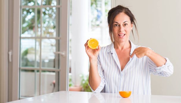 Frau mittleren Alters hält orangefarbene Früchte mit überraschendem Gesicht und zeigt mit dem Finger auf sich selbst - Foto, Bild