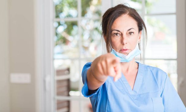 Μέσης ηλικίας γυναίκα γιατρός, δείχνοντας με το δάχτυλο στη φωτογραφική μηχανή και σε εσάς, να το χέρι συνδεθείτε, θετική και σίγουρη κίνηση από το μέτωπο - Φωτογραφία, εικόνα