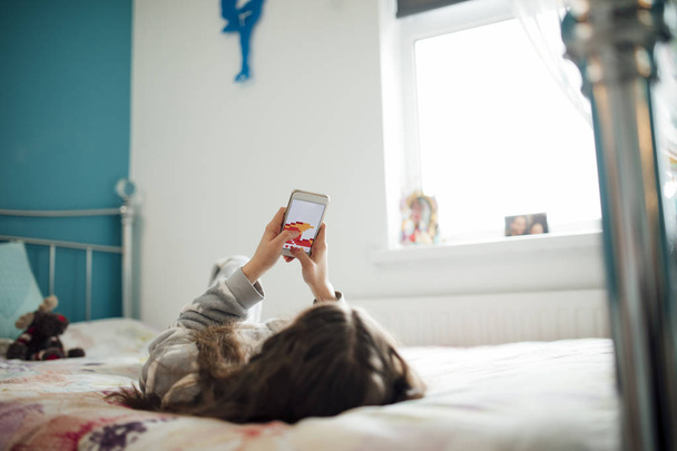 Petite fille est allongée sur son lit, jouant à un jeu sur son smartphone
. - Photo, image