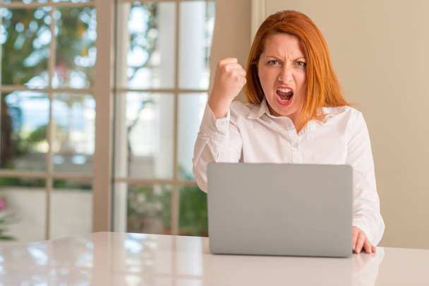 Mujer pelirroja usando computadora portátil en casa molesto y frustrado gritando con ira, loco y gritando con la mano levantada, concepto de ira
 - Foto, Imagen