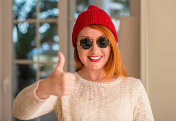Elegante donna rossa che indossa berretto di lana e occhiali da sole felice con grande sorriso facendo segno ok, pollice in su con le dita, segno eccellente
 - Foto, immagini