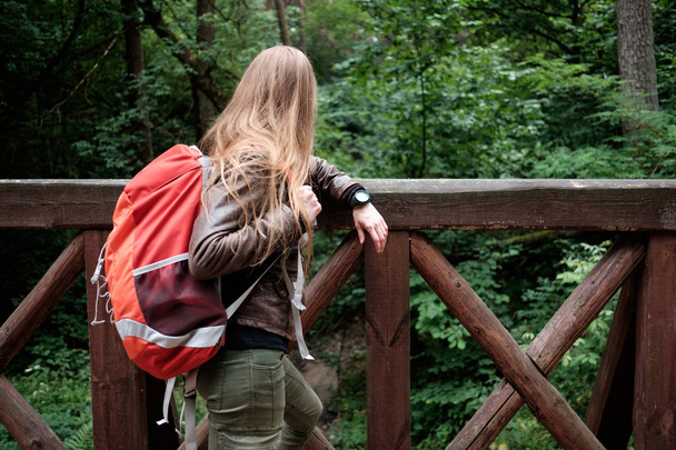 Jeune femme rousse aux cheveux longs voyage avec un sac à dos de voyage rouge, se repose sur un pont en bois sur la rivière de la forêt et regarde loin. Concept voyage
. - Photo, image