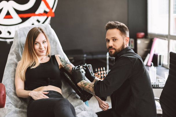 Chica tatuada y tatuador profesional felizmente mirando en cámara mientras hace tatuaje a mano por máquina de tatuaje en estudio
 - Foto, Imagen