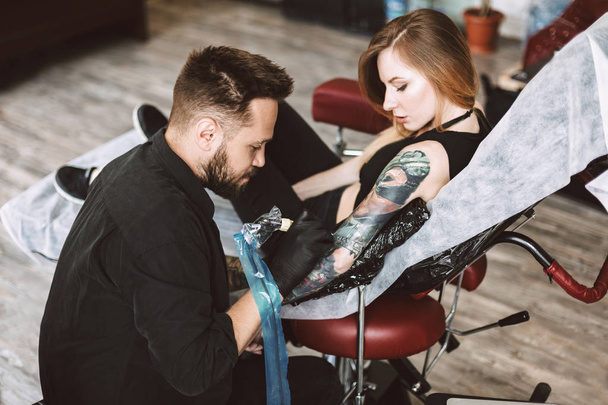 Ammattimainen tatuoija tekee tatuoinnin käsillä tatuointikoneella, kun taas tyttö harkitsee katsellen prosessia modernissa studiossa.
 - Valokuva, kuva