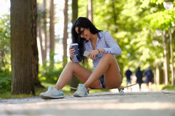 Riendo chica morena sexy en gafas con una camisa y pantalones cortos sentados en un monopatín y sostiene una taza de café para llevar y teléfono inteligente en un parque
 - Foto, imagen