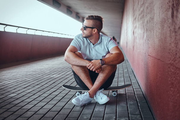 Portrait of a handsome fashionable skater guy in sunglasses dressed in a white shirt and shorts sitting on a skateboard under the bridge, looking away. - Foto, immagini