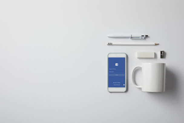 top view of smartphone with facebook app and various supplies on white tabletop - Fotografie, Obrázek