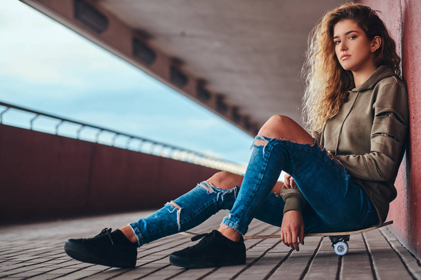 Portrait of a young woman dressed in a hoodie and ripped jeans leaning on a wall while sitting on a skateboard at bridge footway. - Photo, image