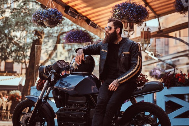 Motociclista elegante em óculos de sol vestido com uma jaqueta de couro preto, sentado em sua motocicleta retro feita sob medida perto do terraço de um café
. - Foto, Imagem