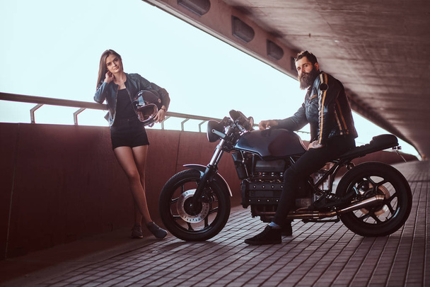 Portrait of an attractive couple - brutal bearded biker in black leather jacket sitting on a motorcycle and his young sensual brunette girlfriend on a footway under a bridge, looking at a camera. - Φωτογραφία, εικόνα
