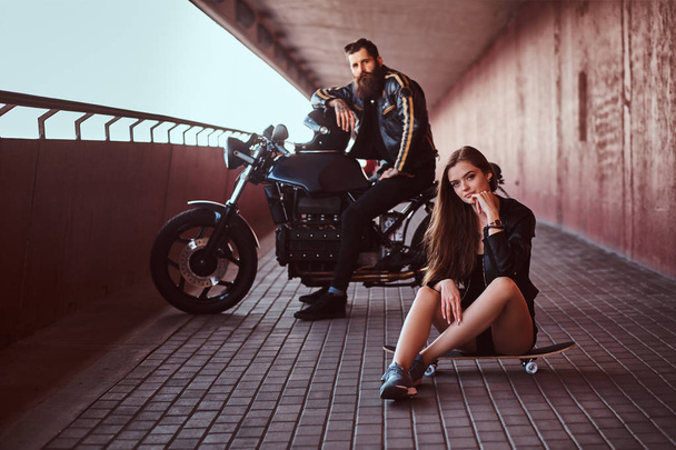 Πορτρέτο του ένα αισθησιακό μελαχρινή κοπέλα κάθεται σε ένα skateboard και βάναυση γενειοφόρος ποδηλάτης μαύρο δερμάτινο σακάκι κάθεται σε μια μοτοσικλέτα σε ένα μονοπάτι κάτω από μια γέφυρα, βλέπουν μια φωτογραφική μηχανή. - Φωτογραφία, εικόνα