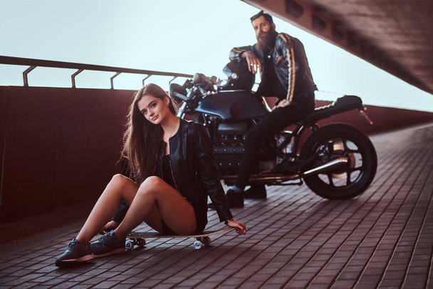 Portrait of a young sensual brunette girl sitting on a skateboard and brutal bearded biker in black leather jacket sitting on a motorcycle on a footway under a bridge, looking at a camera. - Foto, afbeelding