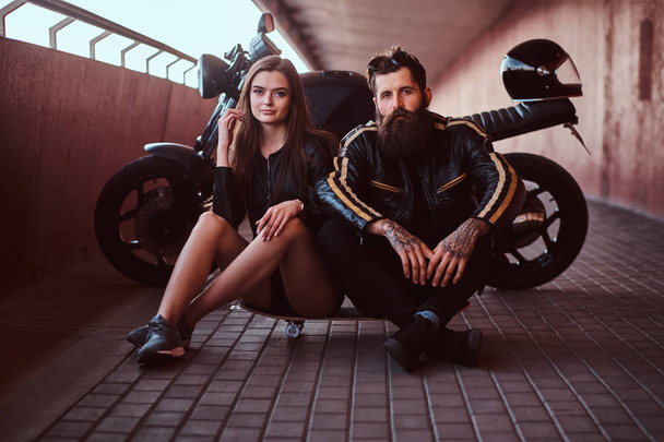 Brutal bearded biker in black leather jacket and sensual brunette girl sitting together on a skateboard near custom-made retro motorcycle on a footway under bridge. - Photo, Image
