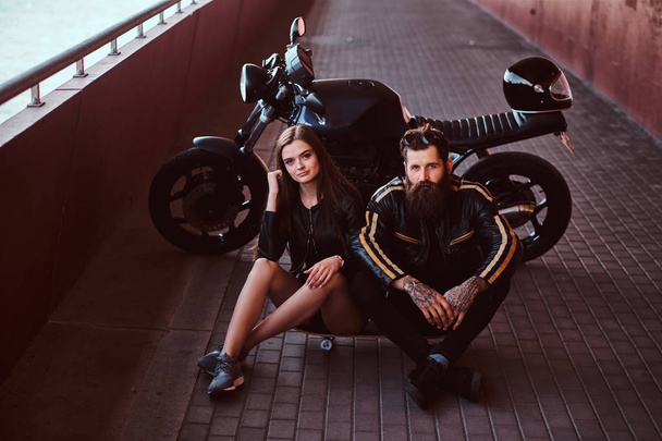 Brutal bearded biker in black leather jacket and sensual brunette girl sitting together on a skateboard near custom-made retro motorcycle on a footway under bridge. - Photo, Image