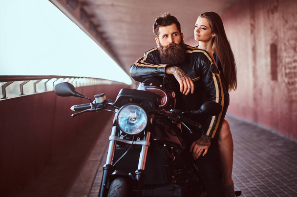 Brutal vousatých biker v černé kožené bundě a smyslná tmavovláska, sedí spolu na zakázku retro motocykl na pěší pod mostem. - Fotografie, Obrázek