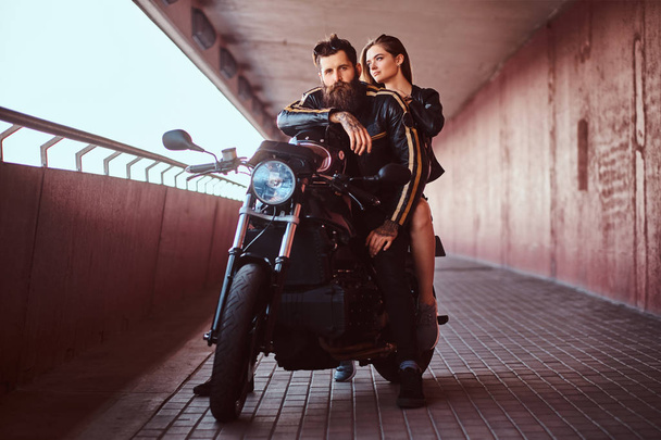 Brutaali parrakas moottoripyöräilijä mustassa nahkatakissa ja aistillinen brunette tyttö istuu yhdessä mittatilaustyönä tehdyllä retro-moottoripyörällä kävelyllä sillan alla
. - Valokuva, kuva