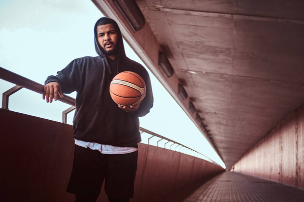Портрет темнокожего бородатого парня в черной толстовке и спортивных шортах, прислонившегося к ограде, стоя рядом с баскетболом
. - Фото, изображение