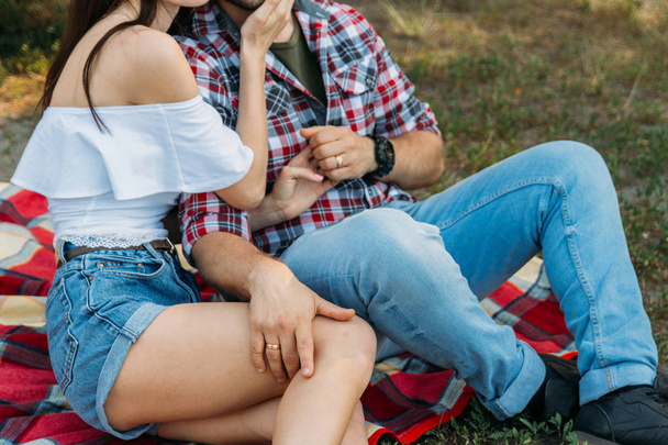 a guy and a girl are sitting on a plaid veil on the grass, hugging and kissing. a man in a plaid shirt and jeans,with a watch on his arm, a girl dressed in a white blouse and denim shorts.holding hands. the man put his hand on the girl's foot.sexual  - Zdjęcie, obraz