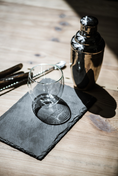 close-up view of shiny empty glass on slate board, shaker and tongs on wooden table - Photo, Image