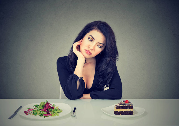Young woman sitting at table with salad and cake craving for dessert and having diet dilemma on gray background - Φωτογραφία, εικόνα