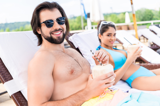 young couple in sunglasses relaxing on sunbeds with coconut cocktails - Foto, imagen