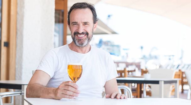 Handsome senior man drinking beer at restaurant with a happy face standing and smiling with a confident smile showing teeth - Photo, image