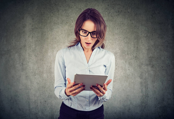 junge schlanke Frau mit Brille schaut erstaunt auf Laptop, während sie Nachrichten auf grauem Hintergrund liest - Foto, Bild