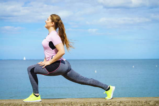 Super fit attractive young woman wearing fashionable outfit working out being active outside during sunny weather. Stretching her legs or practice yoga next to sea - Photo, image