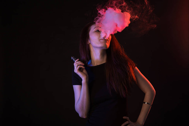 Jonge vrouw in neon licht en rook van e-sigaretten of vape op zwarte achtergrond - Foto, afbeelding