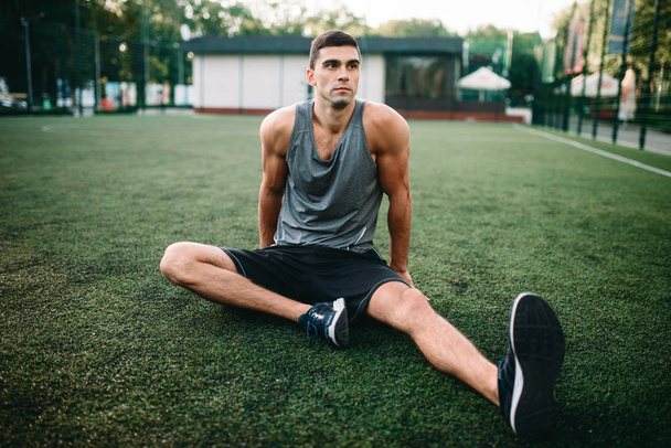 Male athlete on outdoor fitness workout. Sportsman sits on grass and doing stretching exercise - Foto, imagen