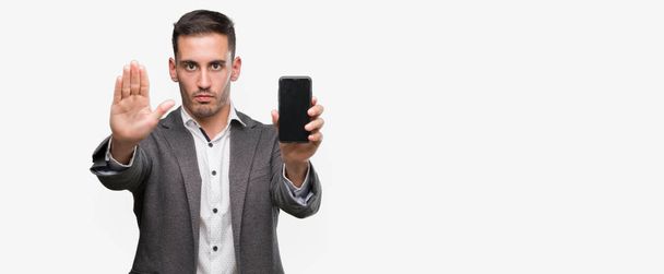 Όμορφος νεαρός άνδρας, χρησιμοποιώντας το smartphone με το ανοιχτό χέρι κάνει στοπ με σοβαρές και αυτοπεποίθηση έκφραση, χειρονομία άμυνα - Φωτογραφία, εικόνα
