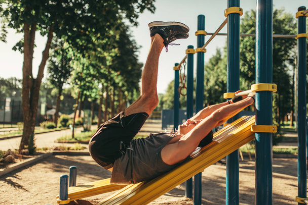 Hombre en ropa deportiva haciendo ejercicios abdominales usando barra horizontal, entrenamiento de fitness al aire libre. Deportista fuerte en el entrenamiento deportivo en el parque
 - Foto, imagen