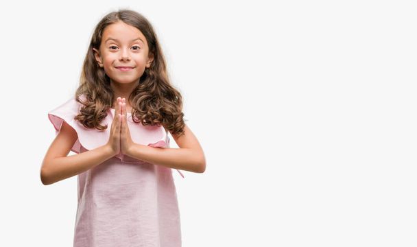 Brunette hispanic girl wearing pink dress praying with hands together asking for forgiveness smiling confident. - Photo, image