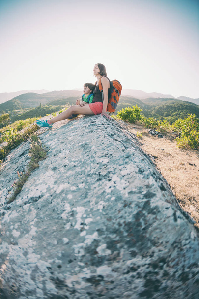 A woman is traveling with a child. Mother with a son is sitting on a large rock. Climb to the top of the mountain with children. The boy with the backpack climbed to the top. Active vacations. - Photo, image